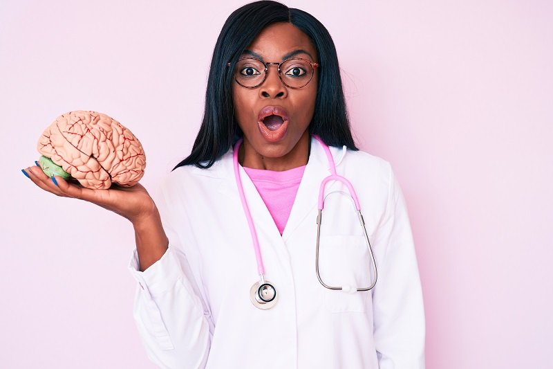 Young african american woman wearing doctor coat holding brain scared and amazed with open mouth for surprise, disbelief face